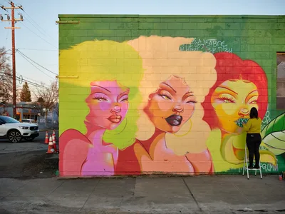 Multidisciplinary artist Villatose applies finishing touches to Jardín Cubana, a striking mural featuring three colorful female portraits on a green cinderblock wall in Portland's Alberta Arts District. The artist, positioned on a small stepladder, works with concentrated focus while the late afternoon light illuminates the vivid yellow, pink, and coral faces that emerge from the urban canvas. Traffic cones and power lines frame this moment of artistic creation in the heart of Northeast Portland's creative corridor.