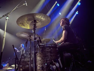 Julian Dorio commands the stage behind his drum kit during Band of Skulls' 10 Year Anniversary Tour at Webster Hall in Manhattan. Theatrical blue stage lights pierce through atmospheric haze, creating dramatic shafts of illumination that frame the focused drummer in profile. The intimate venue setting captures the raw energy of live performance, with gleaming cymbals suspended overhead and a bassist visible in the background, all enveloped in the moody ambiance of concert lighting.