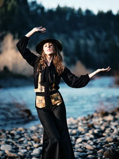 A woman in flowing black attire and wide-brimmed hat strikes an expressive dance pose along the rocky riverbed of Multnomah County's Floodplain Trail. Golden hour light illuminates her auburn hair and ornate vest details while she moves gracefully against the backdrop of the Columbia River and distant evergreen forests. The composition captures a moment of spontaneous movement and connection with Oregon's natural landscape.