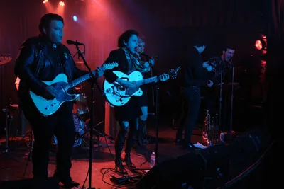Victor Franco, Charlotte McCaslin, and Faith Grossnicklaus of Roselit Bone perform under saturated red and blue stage lighting at Dante's in Portland, Oregon during RX Fest 2019. The intimate venue's moody atmosphere envelops the trio as Franco commands the microphone on the left, McCaslin centers with her hollow-body guitar, and Grossnicklaus anchors the right side. Theatrical lighting cuts through the smoky darkness, casting the musicians in electric hues while cables snake across the cluttered stage floor.