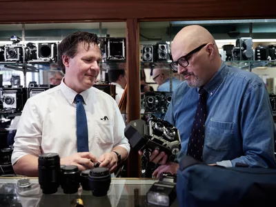 Mike Knight and Arthur Ruckle examine vintage cameras with practiced expertise inside Blue Moon Camera & Machine on North Lombard Street in Portland's Saint Johns neighborhood. Knight, wearing a crisp white shirt and blue tie, handles camera lenses while Ruckle, distinguished in a denim shirt and glasses, inspects a classic camera body. The warm interior lighting illuminates shelves lined with an impressive collection of vintage cameras and equipment, creating an atmosphere of craftsmanship and mechanical precision. Their focused collaboration speaks to decades of shared passion for analog photography and the careful curation that defines this beloved Portland institution.