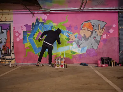 A lone muralist works intently on a vibrant wall painting in the industrial setting of Clay Creative's parking garage in Southeast Portland. The artist, dressed in black, adds details to a fantastical composition featuring geometric shapes, graffiti-style letters, and a whimsical character in a hoodie against a bold magenta background. Spray paint cans and art supplies scatter across the concrete floor, while exposed ceiling infrastructure frames this underground creative sanctuary.