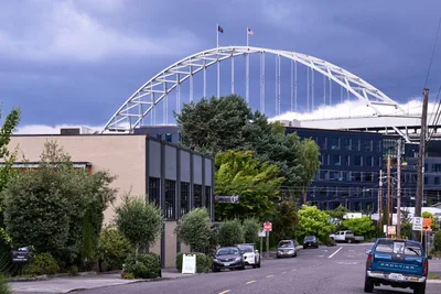 The iconic white steel arch of Portland's Fremont Bridge soars majestically against a dramatic sky of periwinkle and storm clouds, its suspension cables creating rhythmic vertical lines. Viewed from the tree-lined streets of the Slab Town neighborhood, the modernist bridge dominates the urban landscape while leafy deciduous trees and contemporary buildings frame the foreground. Two flags flutter atop the bridge's peak, while parked cars and residential streets below emphasize the human scale against this monumental infrastructure.
