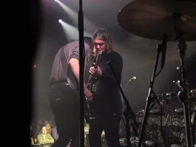 Emma Richardson and Russell Marsden of Band of Skulls perform during their 10 Year Anniversary Tour at Manhattan's historic Webster Hall. Dramatic stage lighting cuts through the smoky atmosphere as Richardson grips her guitar neck while Marsden leans in beside her, both musicians silhouetted against the venue's iconic backdrop. The intimate concert setting captures the raw energy of their decade-spanning musical partnership, with drum kit and amplifiers forming the stage's industrial landscape.
