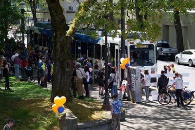 Crowds gather beneath the dappled shade of mature trees along Lloyd Center's sidewalk to witness the retirement of Portland's iconic MAX Type 1 train on a bright April afternoon. Orange and blue balloons punctuate the celebratory scene as transit enthusiasts, families, and community members line the platform area, their shadows creating intricate patterns across the concrete. The white articulated train car displays its familiar blue destination sign while people of all ages document this historic moment in Portland's transportation legacy.