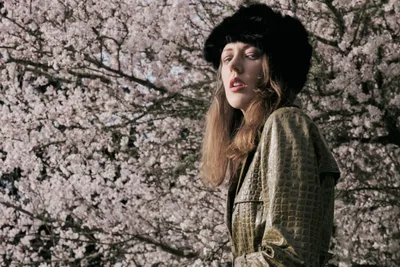 Model Rhianna Atwood stands contemplatively beneath a canopy of delicate cherry blossoms in full bloom at Portland's Mount Tabor park. Dappled natural light filters through the pale pink petals, illuminating her serene expression as she gazes upward while wearing a luxurious olive quilted coat and dramatic black fur hat. The spring hanami scene captures a moment of quiet reflection amid nature's ephemeral beauty, with the abundant sakura blossoms creating an ethereal backdrop that speaks to seasonal renewal and peaceful introspection.