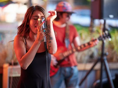 Aubrey Gordon leans into a vintage-style microphone with passionate intensity, her auburn hair catching golden light as she performs at Portland's bustling Mercado. The tattooed vocalist in a sleek black top commands attention while a guitarist in red flannel provides melodic accompaniment in the softly blurred background. Warm evening light bathes the outdoor venue, creating an intimate atmosphere that bridges the energy between performer and audience in Southeast Portland's vibrant cultural hub.