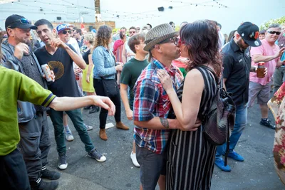A couple shares a tender kiss in the center of a bustling outdoor day party at White Owl Social Club in Portland. The man in a plaid shirt and tweed cap embraces a woman in pinstripe while dozens of festival-goers mill around them on the asphalt courtyard. String lights overhead and the casual summer attire of attendees create an intimate contrast between private romance and public celebration during this Your Sunday Best season opener.