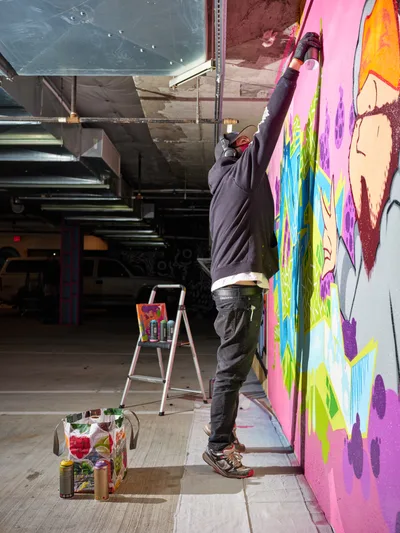 A masked street artist stretches upward to paint a kaleidoscopic mural featuring anime-inspired characters in brilliant pinks, purples, and greens on the concrete wall of Clay Creative's parking garage in Southeast Portland. The industrial space, with its exposed ceiling and metal framework, provides a raw canvas for the emerging artwork. Spray paint cans and art supplies scattered on the concrete floor tell the story of creation in progress, while natural light filters through the urban space.