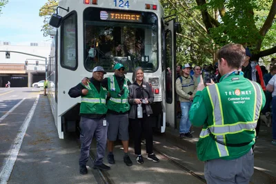 TriMet employees in vibrant green safety vests gather beside the white Type 1 MAX train numbered 124 during its retirement ceremony in Portland's Lloyd District. The scene captures a bittersweet moment as transit workers pose for commemorative photos, their camaraderie evident against the backdrop of leafy street trees and urban infrastructure. Dappled sunlight filters through the canopy, creating a nostalgic atmosphere as onlookers witness the end of an era for Portland's pioneering light rail system.