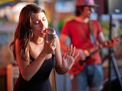 Vocalist Aubrey Gordon delivers an impassioned performance into a vintage-style microphone at Portland Mercado, her eyes closed in musical concentration as golden hour light bathes her expressive face. Her raised hand gestures emphasize the emotional intensity of her song while a blurred musician in red provides atmospheric depth in the background. The Southeast Portland food hall's colorful storefronts create a vibrant bokeh backdrop, capturing the intimate energy of live music in this multicultural community gathering space.