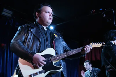 Victor Franco of Roselit Bone commands the dimly lit stage at Dante's in Portland, his cream-colored Telecaster gleaming under moody blue stage lights. Clad in a weathered black leather jacket adorned with pins, Franco's concentrated expression captures the raw energy of underground rock performance. The atmospheric lighting carves dramatic shadows across his face while a bandmate remains partially visible in the background, creating an intimate yet electrifying concert scene.