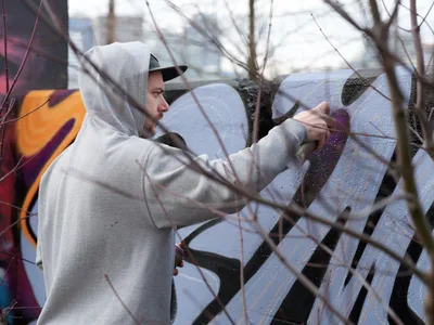 Travis Fields, known as Campographic, carefully applies purple paint to a concrete wall at the Clay Creative Building in southeast Portland's Hosford-Abernethy neighborhood. The hooded street artist works methodically behind bare winter branches that create natural framing across the composition. His gray hoodie contrasts against the textured concrete surface as vibrant orange and purple graffiti elements emerge in the industrial creative space.