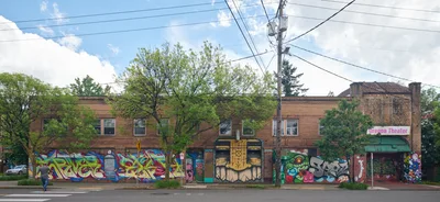 Vibrant murals cascade across the brick facade of the former Oregon Theater building on Southeast Division Street in Portland's Richmond neighborhood. The colorful street art, featuring bold graffiti-style lettering, masked figures, and abstract designs, transforms this historic two-story commercial structure into a living canvas. A lone pedestrian crosses the quiet street under overcast spring skies, while power lines create geometric patterns against the cloudy backdrop. The murals, sponsored by the Portland Street Art Alliance, breathe new life into this cultural landmark along one of the city's most dynamic corridors.