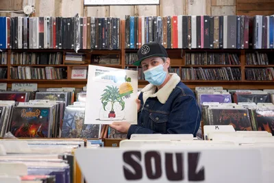 A masked record store employee in a sherpa-lined denim jacket and black cap proudly displays a vinyl album featuring botanical artwork against the towering backdrop of Music Millennium's legendary collection during Record Store Day 2022. The warm, diffused lighting illuminates countless albums organized across wooden shelves that stretch from floor to ceiling, creating an intimate portrait of vinyl culture in Portland's iconic Kerns neighborhood. The scene captures the enduring passion for physical music media during this annual celebration of independent record stores, with alphabetized sections and genre dividers creating a maze of musical discovery.
