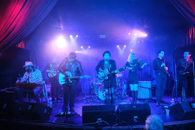 Six musicians command the intimate stage at Dante's in Portland, bathed in electric blue and magenta stage lighting that transforms the venue into a nocturnal dreamscape. The lead vocalist grips a hollow-body guitar while a violinist adds ethereal textures, flanked by keyboards, bass, and drums in this captivating indie ensemble. Crimson curtains frame the performance space as dramatic spotlights slice through the atmospheric haze, creating an otherworldly concert experience.