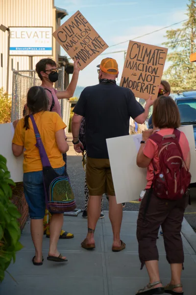 A small group of climate activists gathers on a sun-drenched sidewalk outside Representative Kurt Schrader's office in Oregon City, Oregon, holding handmade cardboard signs demanding climate action over moderation. The golden afternoon light illuminates the diverse group of protesters, including families with children carrying colorful backpacks, as they position themselves near the building's entrance. The scene captures grassroots political engagement in a suburban Pacific Northwest setting, with the activists' casual summer attire and determined postures reflecting the urgency of their environmental message.