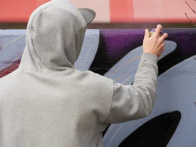 Travis Fields, known as Campographic, works intently on a colorful mural at the Clay Creative Building in Southeast Portland's Hosford-Abernethy neighborhood. Wearing a gray hoodie and cap, the artist applies purple and black paint with focused precision, his outstretched hand guiding the brush across the wall. The warm coral-pink background suggests late afternoon light filtering through the creative space, capturing a moment of artistic dedication in one of Portland's vibrant cultural hubs.