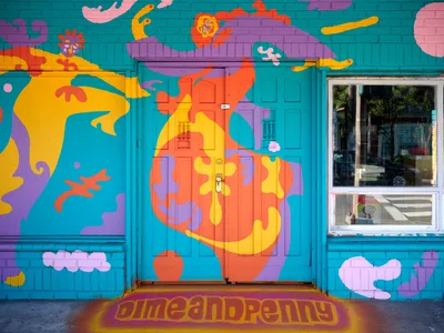 A vibrant street mural by artist Naomi Likayi transforms a turquoise building facade in Portland's Alberta neighborhood into a fantastical wonderland. Fluid organic shapes in coral, yellow, and purple dance across the wall, featuring playful cloud formations and flowing abstract elements that seem to melt and merge into one another. The psychedelic composition frames a central doorway and window, creating an immersive portal effect that invites passersby into this colorful urban dreamscape.