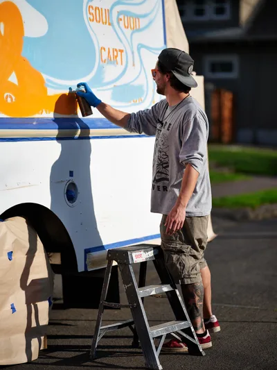 A dedicated muralist in casual work attire and backward cap applies spray paint to the vibrant exterior of a white food truck on a quiet Portland street. The artist works methodically from a metal stepladder, adding finishing touches to colorful graphics that include orange and blue swirls alongside 'SOUL FOOD CART' lettering. Golden afternoon light bathes the scene, casting crisp shadows across the asphalt while residential buildings blur softly in the background.