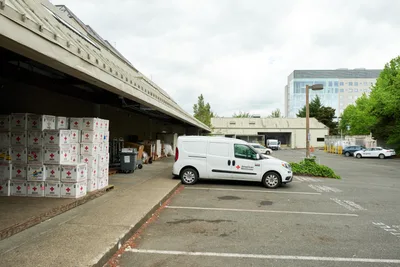 Under overcast Pacific Northwest skies, a white American Red Cross delivery van sits positioned at a concrete loading dock in the organization's Cascades Region facility on North Vancouver Avenue. Towering stacks of empty blood platelet boxes marked with red crosses await collection inside the shadowed warehouse bay, while the modern glass facade of a Portland office building rises in the background. The utilitarian scene captures the operational backbone of humanitarian aid distribution in Oregon's largest city.