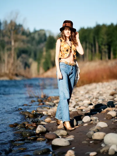 A young woman in vintage-inspired attire poses confidently along the rocky shoreline of the Columbia River near the Floodplain Trail in Multnomah County. Bathed in warm golden hour light, she wears a floral crop top, high-waisted jeans, and a dark fedora while standing among water-smoothed stones. The soft focus background reveals the river's gentle current and dense Pacific Northwest forest, creating an intimate portrait that captures both personal style and Oregon's natural beauty.