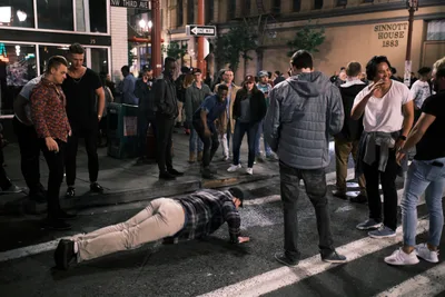 A late-night street scene at the corner of NW Third Avenue in Portland's Chinatown as bars and clubs close for the night. A young man in a plaid shirt and khaki pants does push-ups in the middle of the wet street while a crowd of onlookers gathers around him, some laughing and watching with amusement. The historic Sinnott House building from 1883 is visible in the background, along with a one-way street sign and the glow of bar lights on the rain-slicked sidewalk.