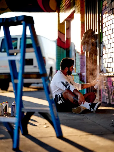 Muralist Heysus sits cross-legged on the sidewalk, painting a vibrant mural on a white-tiled wall in Portland's Alberta neighborhood. Golden hour light bathes the scene, casting long shadows from a blue metal ladder positioned nearby. Spray paint cans scattered on the ground reveal his working process, while the artist focuses intently on adding colorful details to the emerging artwork at the intersection of Killingsworth and Northeast 15th Avenue.