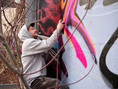 Travis Fields, known as Campographic, applies vibrant magenta and orange paint to a concrete wall at the Clay Creative Building in southeast Portland's Hosford-Abernethy neighborhood. Crouched behind protective metal fencing, the hooded artist methodically works on his mural while winter-bare branches frame the scene. The industrial setting and natural lighting create an authentic documentary atmosphere capturing the intersection of urban art and creative community spaces.