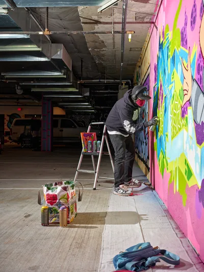 A hooded artist in dark clothing meticulously works on a large-scale mural featuring explosive bursts of pink, yellow, blue and purple paint across the concrete wall of a converted parking garage. The industrial space at Clay Creative in Southeast Portland's Hosford-Abernethy neighborhood buzzes with creative energy as exposed ceiling infrastructure frames the scene. Spray paint cans, brushes, and art supplies scatter across the polished concrete floor, while natural light filters through the urban warehouse setting, illuminating the transformation of raw space into artistic canvas.