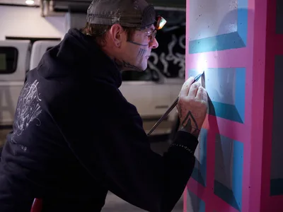 Artist Rabbitt meticulously applies precise brushstrokes to a vibrant geometric mural within the industrial confines of Portland's Clay Creative building parking garage. His tattooed hands work with steady concentration as magenta and cyan geometric forms emerge on the concrete wall, illuminated by a focused headlamp that cuts through the garage's dim atmosphere. The juxtaposition of raw urban infrastructure and carefully crafted geometric patterns captures the essence of Portland's underground art scene.