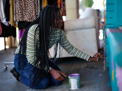 Naomi Likayi sits on the concrete sidewalk, carefully applying her signature to a vibrant turquoise wall mural in Portland's Alberta Arts District. Her long braided hair cascades over a striped shirt as she leans forward with focused concentration, paintbrush in hand and paint can beside her. The scene captures an intimate moment of artistic completion against the backdrop of the neighborhood's characteristic colorful storefronts and creative energy.