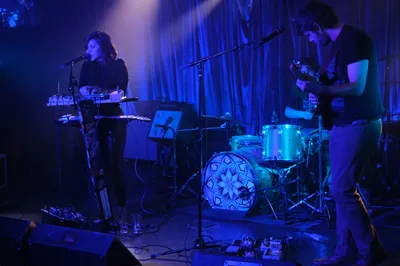 Experimental band Sun Blood Stories performs in the intimate, moody confines of Dante's in downtown Portland during Rx Fest. Electric blue stage lighting bathes the duo as the keyboardist leans into the microphone while the drummer maintains rhythm behind a geometric-patterned kit. The atmospheric lighting creates deep shadows against flowing curtains, capturing the mesmerizing sound of the Boise-founded band now based in Portland.