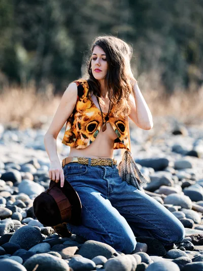 A young woman sits contemplatively among smooth river stones along Oregon's Floodplain Trail, her chestnut hair catching golden afternoon light. She wears a striking animal-print crop top paired with high-waisted denim and layered accessories, embodying free-spirited bohemian style. The shallow depth of field isolates her against the softly blurred forest backdrop, creating an intimate portrait that speaks to wanderlust and connection with nature.