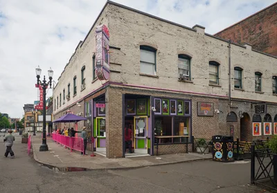 The iconic Voodoo Doughnut shop anchors a weathered brick corner in Portland's Old Town Historic District, its signature purple and green storefront radiating defiant vibrancy against overcast skies. Pink safety barriers channel a socially distanced queue of masked customers along the sidewalk, while the establishment's eclectic signage and window displays maintain their characteristic irreverent charm. The scene captures urban resilience during the pandemic, with pedestrians navigating the altered streetscape beneath heavy Pacific Northwest clouds.