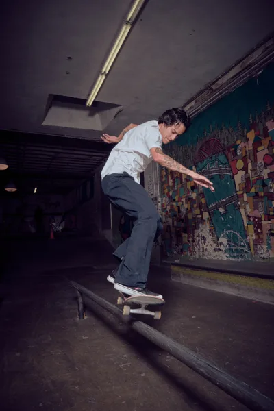 A skateboarder in crisp white shirt and dark pants executes a smooth rail grind in the atmospheric underground space of Commonwealth Skateboarding in Portland's Buckman neighborhood. The moody lighting creates dramatic shadows against weathered concrete walls adorned with vibrant street art murals, while industrial ceiling fixtures cast pools of golden light across the worn wooden rails. The scene captures the raw authenticity of Portland's skateboarding culture within this iconic Southeast 20th Avenue skate shop.