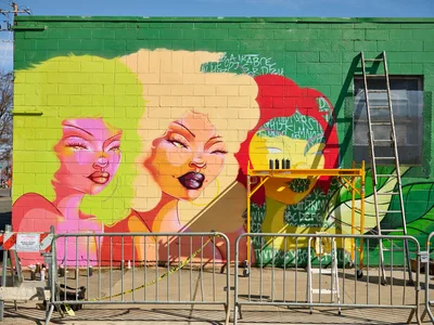 A striking work-in-progress mural titled Jardín Cubana dominates a lime green cinderblock wall along Northeast Alberta Street in Portland's Alberta Arts District. The large-scale artwork features three glamorous female figures rendered in vivid pinks, yellows, and oranges, their faces emerging from swirling colorful hair against the emerald backdrop. Construction scaffolding and safety barriers frame the scene, capturing multidisciplinary artist Vilatose's creative process in this gentrifying neighborhood known for its street art culture.