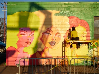 Multidisciplinary artist Vilatose works from a yellow scaffolding platform, adding finishing touches to 'Jardín Cubana,' a striking mural featuring three female figures rendered in brilliant coral, yellow, and green hues on a cinder block wall. The large-scale artwork depicts stylized feminine portraits with flowing hair, painted across the green brick facade in Portland's Alberta Arts District. Golden hour light bathes the scene, casting warm shadows while the artist concentrates on their craft, surrounded by construction barriers and urban infrastructure.