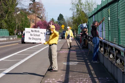 Activists in bright yellow rain gear hold protest signs on North Skidmore Street bridge over Interstate 5 in North Portland during a May Day banner drop action. The central figure displays a white banner reading "No Work No School No Shopping May 1st" while others raise their arms in solidarity. Spring foliage frames the scene as cars pass beneath the overpass, creating a moment of grassroots political theater against the suburban Portland backdrop.