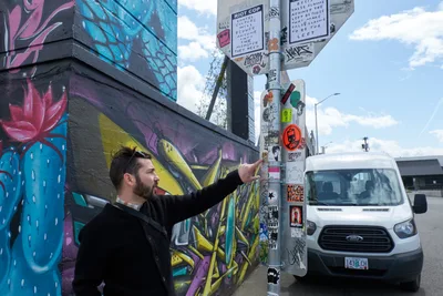 Tomás Valladares of the Portland Street Art Alliance gestures toward a utility pole densely layered with stickers and tags during a guided tour in the Hosford-Abernethy district. The scene unfolds against vibrant graffiti murals featuring blue and pink floral motifs, while a white Ford Transit van waits nearby under an overcast afternoon sky. His animated explanation captures the intersection of grassroots artistic expression and urban culture documentation in Portland's creative corridors.