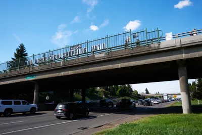 Activists from the Indivisible movement unfurl a protest banner across the Interstate 5 overpass at North Skidmore Street in Portland, Oregon, calling for a May 1st general strike. The white banner with bold black lettering reading 'NO WORK SCHOOL SHOPPING MAY 1st' spans the concrete bridge structure above busy freeway traffic. Against a brilliant blue sky dotted with wispy clouds, the grassroots demonstration transforms mundane urban infrastructure into a platform for political messaging, while commuter traffic flows beneath in the shadowed canyon of the highway corridor.