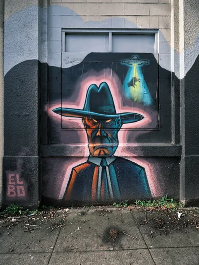 A striking street mural dominates the weathered concrete wall of the former Salvation Army warehouse in Portland's Central Eastside district. The artwork depicts a mysterious figure in a wide-brimmed cowboy hat and suit, rendered in dramatic blues and oranges with a glowing pink aura. Above the figure, a UFO casts an otherworldly teal beam of light, creating a surreal narrative of extraterrestrial encounter. The mural spans the lower portion of the industrial building, with cracked pavement and urban debris at its base, embodying the gritty aesthetic of Portland's commissioned street art scene.