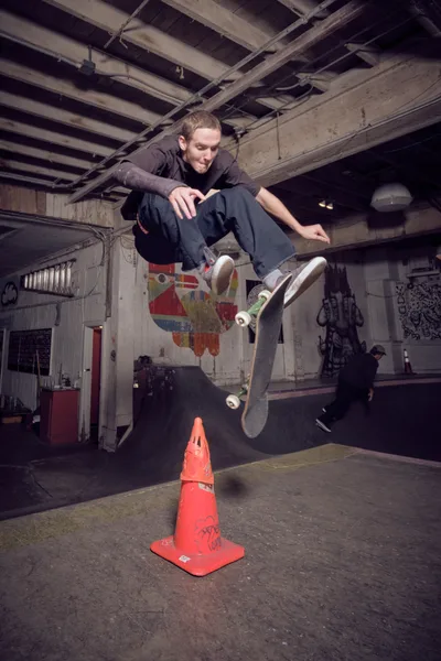 A young skateboarder launches into mid-air above a weathered orange traffic cone at Commonwealth Skateboarding in Portland's Buckman neighborhood. The exposed ceiling beams and graffiti-covered concrete walls create a raw, underground atmosphere, while another skater practices in the shadowed background. Dramatic lighting captures the precise moment of athletic suspension, highlighting the urban skate culture thriving beneath the city streets.