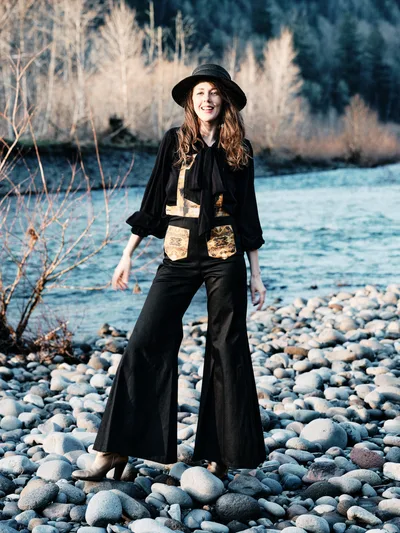 A joyful woman in bohemian attire stands confidently on a pebbled riverbank along the Sandy River in Multnomah County, Oregon. She wears a black wide-brimmed hat, flowing black pants, and a distinctive vest with golden patches over a dark blouse. The late afternoon light illuminates her auburn hair and genuine smile, while dormant willows frame the rushing water behind her. The rocky riverbed stretches endlessly, capturing the serene wilderness of the Pacific Northwest.