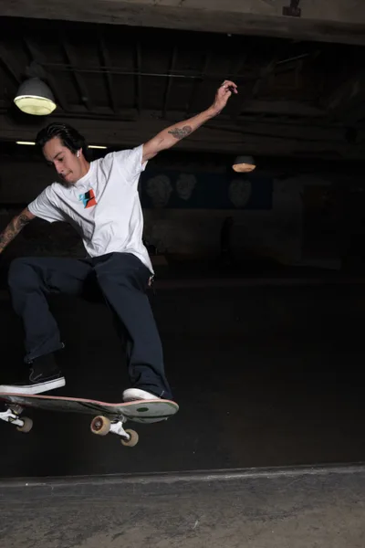 A tattooed skateboarder captures a moment of weightless artistry beneath the industrial ceiling of Commonwealth Skateboarding in Portland's Buckman neighborhood. His extended arm and focused expression reveal the meditative concentration required for technical skateboarding, while the harsh fluorescent lighting creates dramatic shadows against the concrete surfaces. The gritty basement atmosphere of the historic skate shop frames this editorial portrait of urban athleticism and creative expression.