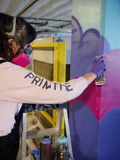 A muralist wearing a pink respirator mask and white sweatshirt emblazoned with 'PRIMITIVE' works on a vibrant purple and magenta mural in the industrial setting of Clay Creative's parking garage in Southeast Portland. Purple latex gloves protect her hands as she wields spray paint cans, their colorful caps arranged on a wooden cart below. The unfinished concrete ceiling and exposed infrastructure frame this creative transformation of utilitarian space into artistic expression.