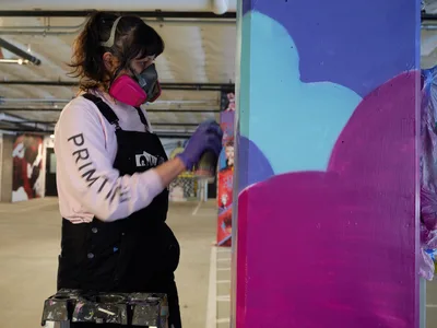 A focused muralist wearing a protective respirator and paint-splattered overalls applies bold magenta and turquoise paint to a large-scale wall piece in the industrial confines of Clay Creative's parking garage. The artist's concentrated brushwork reveals emerging abstract forms against the raw concrete backdrop, while scattered canvases and urban art installations populate the converted creative space in Southeast Portland's Hosford-Abernethy neighborhood.