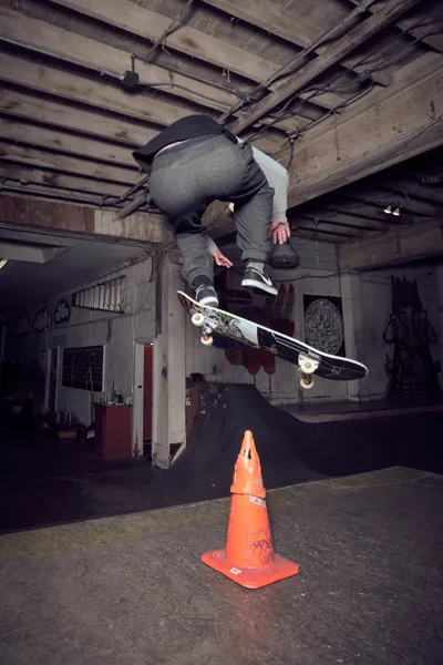A skateboarder launches into a mid-air trick above a weathered orange traffic cone in the raw, industrial interior of Commonwealth Skateboarding in Southeast Portland. The basement space reveals exposed wooden ceiling joists and concrete walls adorned with graffiti, while dramatic shadows cast by overhead lighting create a gritty, underground atmosphere. The athlete, dressed in gray hoodie and dark jeans, demonstrates precise board control as his skateboard floats beneath him in this moment of athletic suspension.