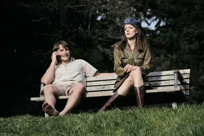 A young woman and young man share a quiet moment on a weathered wooden bench in Mount Tabor Park, Portland. The man on the left speaks into his phone while wearing casual shorts and summer attire, his companion sits poised in military-inspired olive green styling topped with a navy beret. Dappled afternoon light filters through the dense canopy above, casting gentle shadows across the emerald grass beneath their feet.