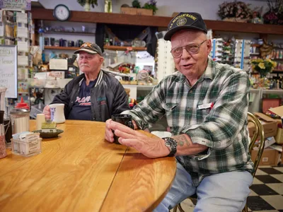 Two elderly regulars, Jewel and Ron, share stories at their corner table in Pattie's Home Plate Café in Portland's Saint Johns neighborhood. Ron, wearing a U.S. Merchant Marine cap and plaid flannel, gestures animatedly while speaking, his weathered hands conveying decades of experience. Jewel sits quietly beside him in a dark jacket, both men surrounded by the warm, cluttered atmosphere of their beloved local diner with its wood shelving lined with knickknacks and memorabilia. The golden wood table between them holds simple white mugs and condiments, testament to countless morning conversations in this community gathering place.